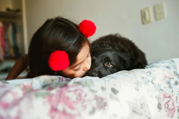 A dark haired young girl, laying next to a black dog, giving the dog a kiss.