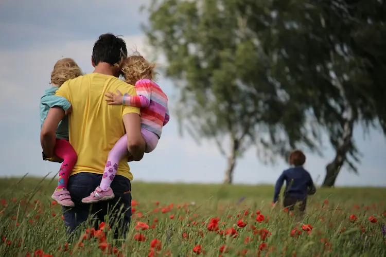 A dad, carrying two kids through a field, another kid running in front of him.