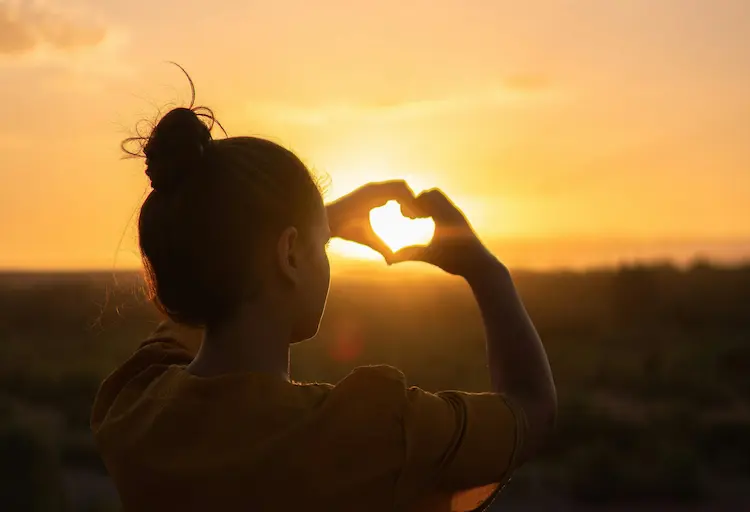 A girl, making a heart with her hands, the sun setting in the background.