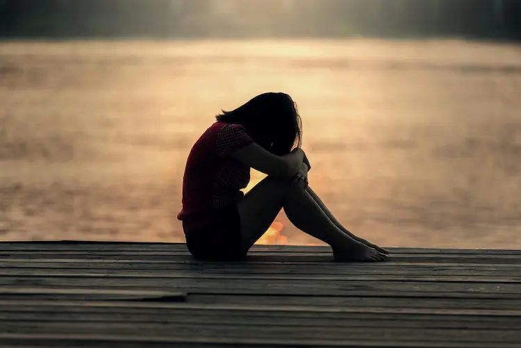 A woman, sitting on a dock by the water, with her head to her knees.